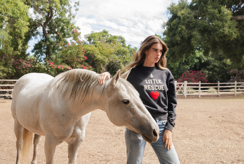 Brunette model standing next to white horse wearing A little Rescue sweatshirt in black