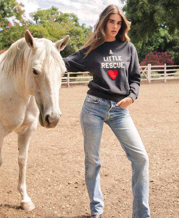 Brunette model standing next to horse wearing A little Rescue sweatshirt in black 