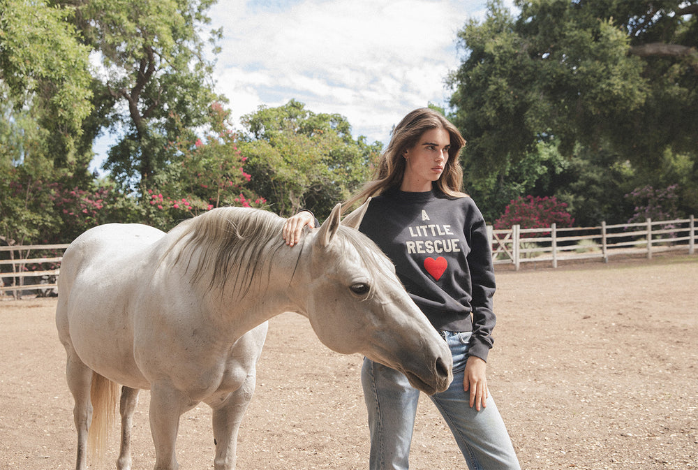 Model wearing the lady & the sailor x A Little Rescue sweatshirt standing next to a white horse