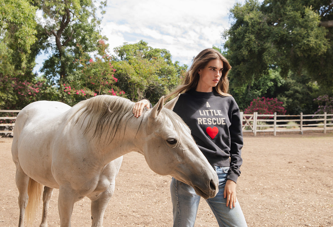 Brunette model standing next to white horse wearing A little Rescue sweatshirt in black